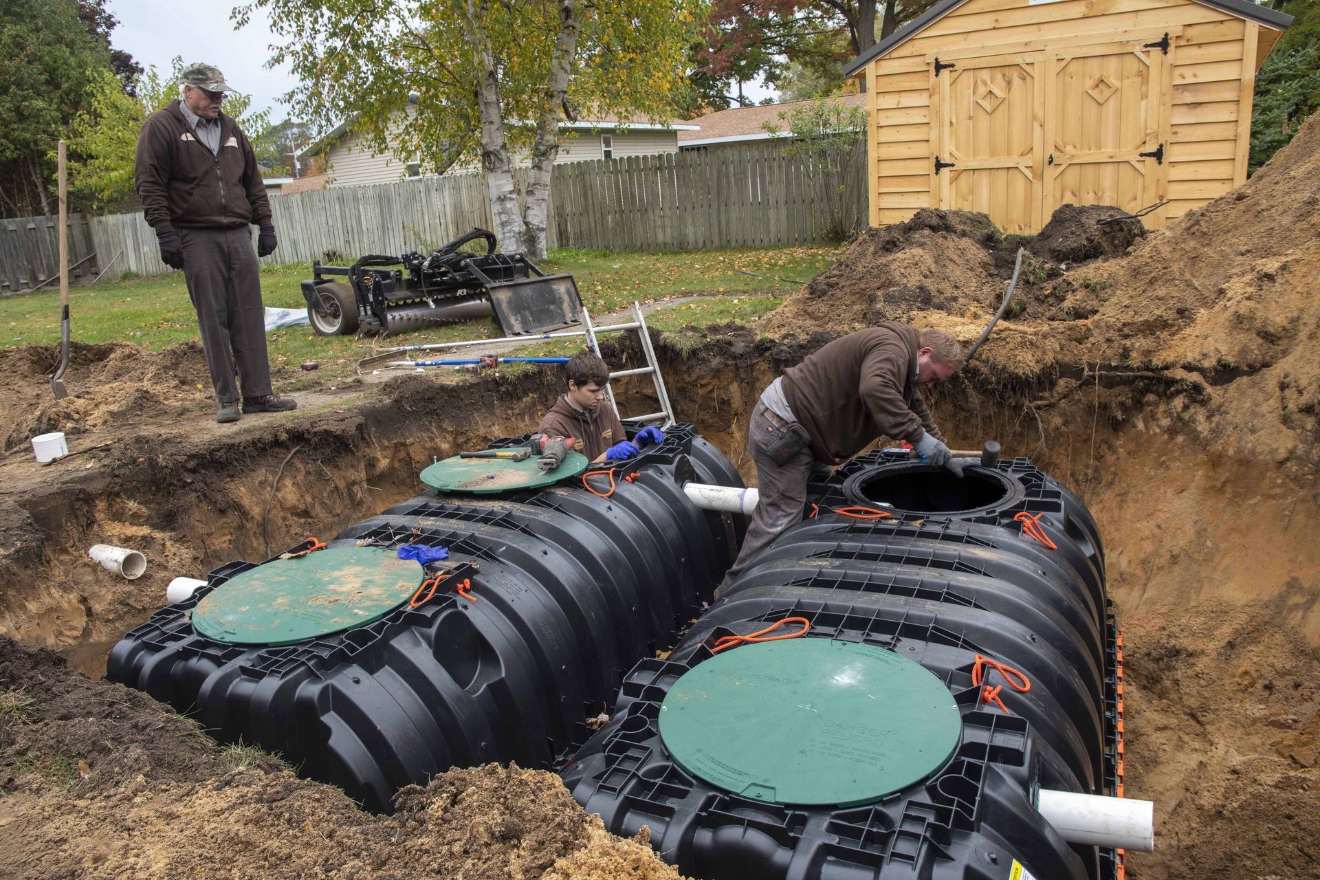 Two workers installing septic tanks in a backyard excavation. One worker inside, the other observes. Green tanks, brown dirt.