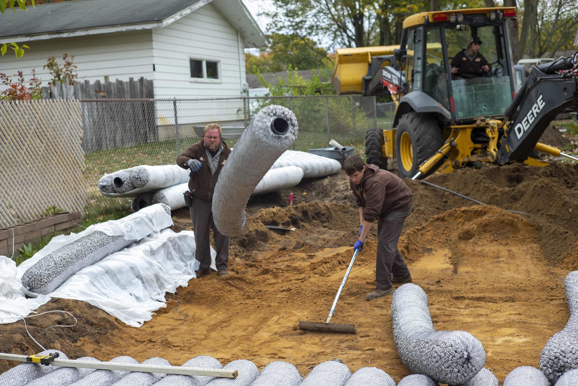 Two people installing drainage system with construction equipment on a residential property.