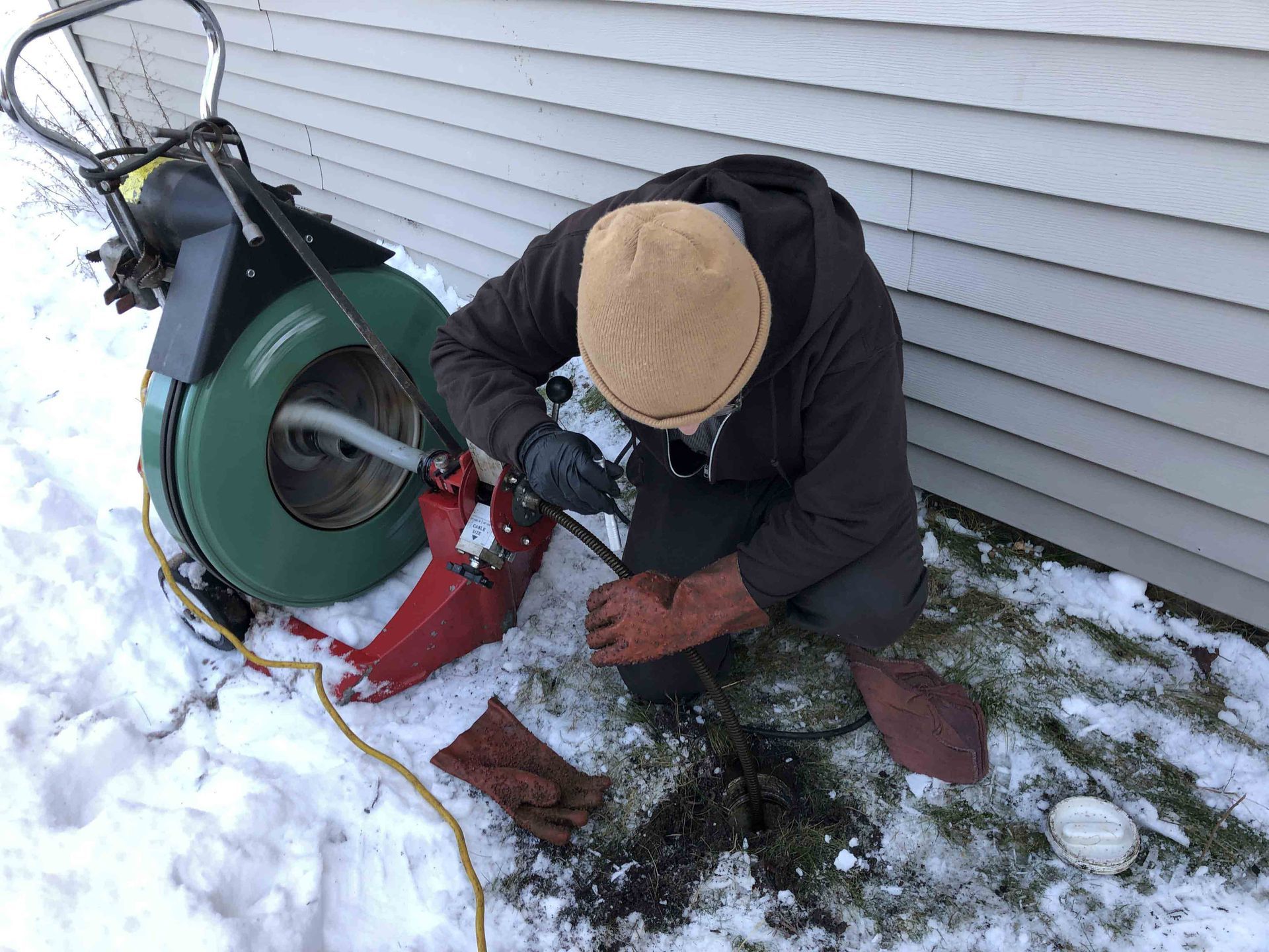 Person unclogging a drain pipe in snowy yard. Using machine, wearing gloves and beanie.