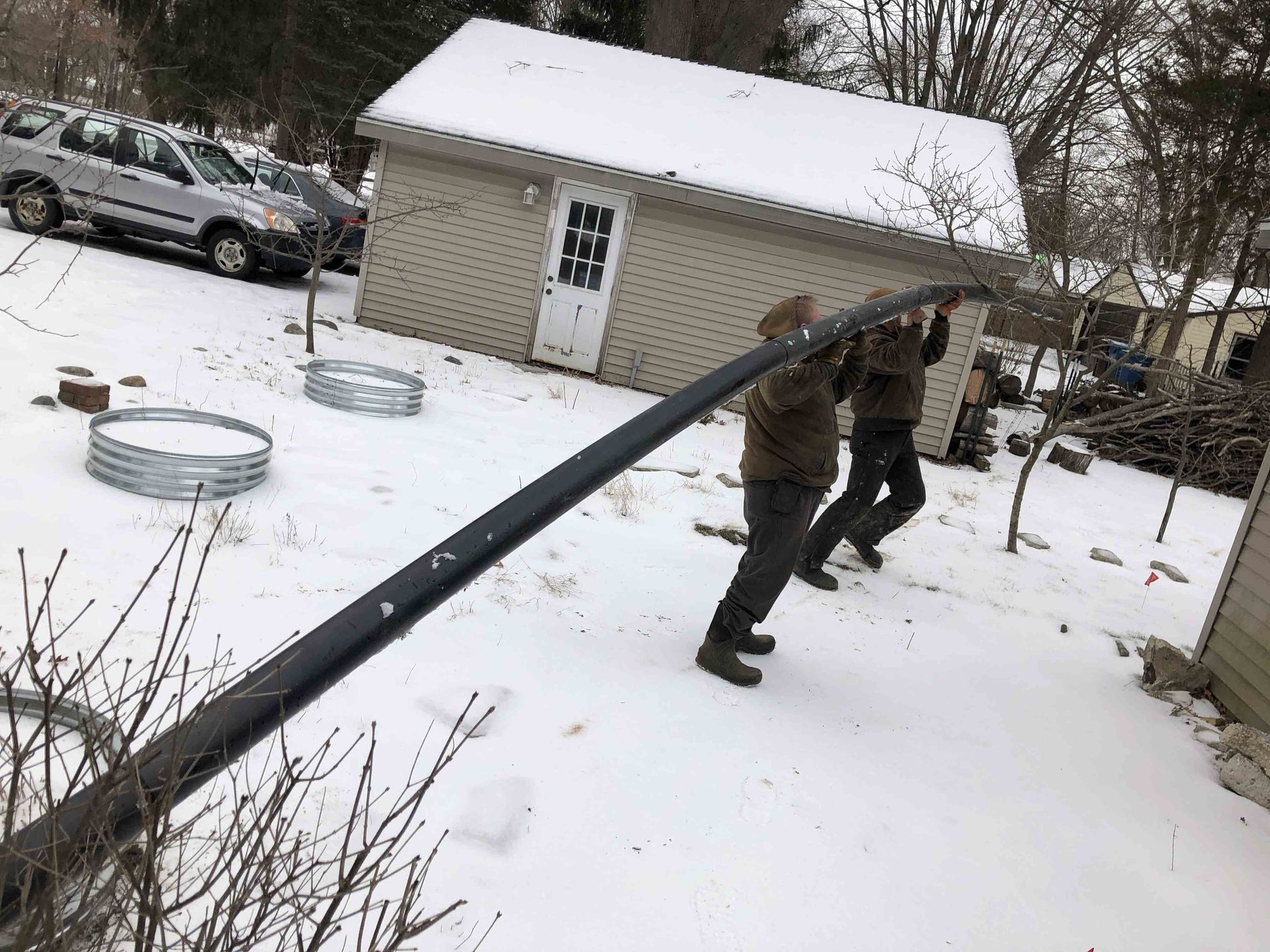 Two people carry a long, black object through a snowy yard, past a shed and cars.
