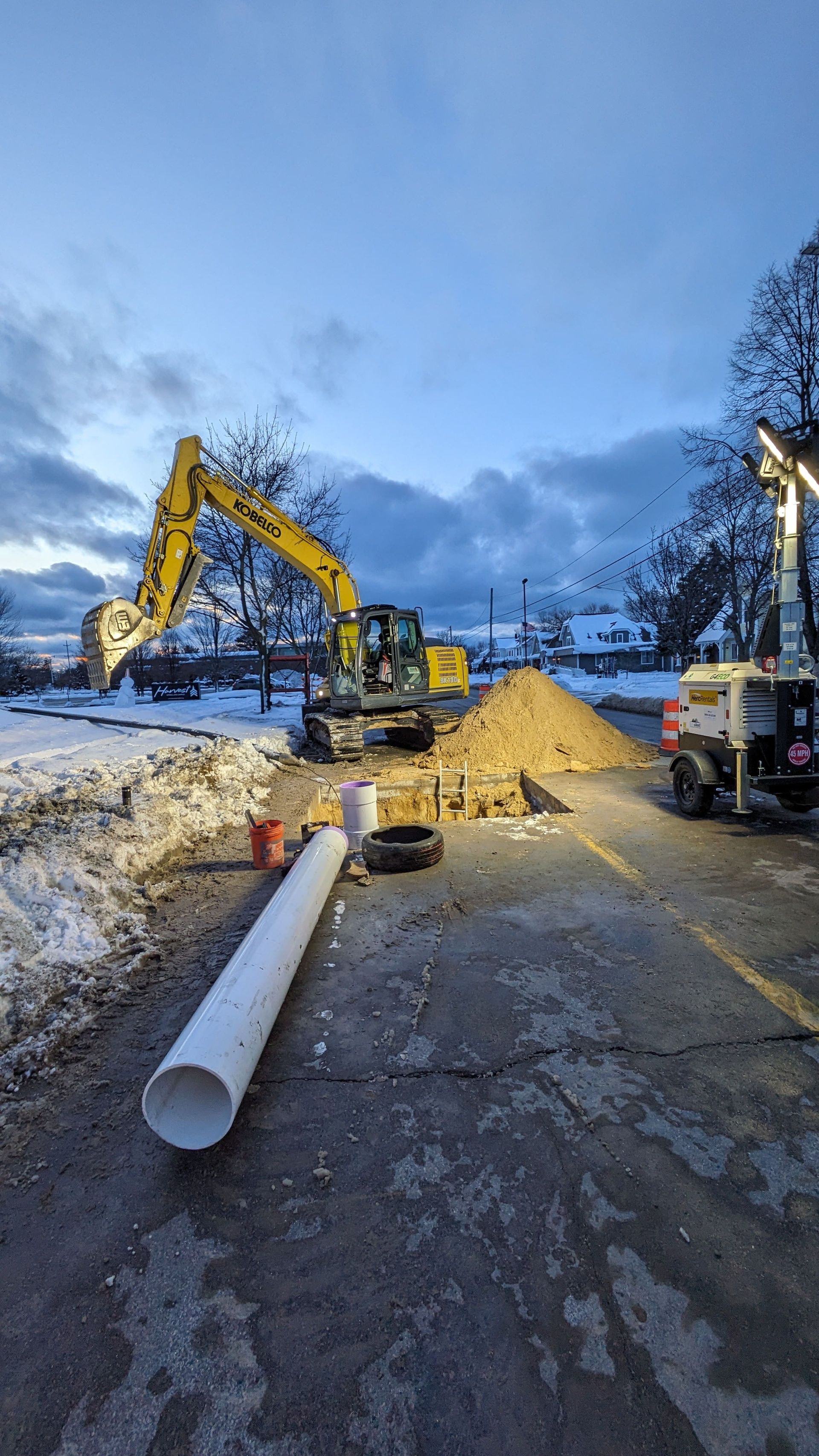Excavator digging near a snowy roadside with large pipe and work lights under a cloudy sky.