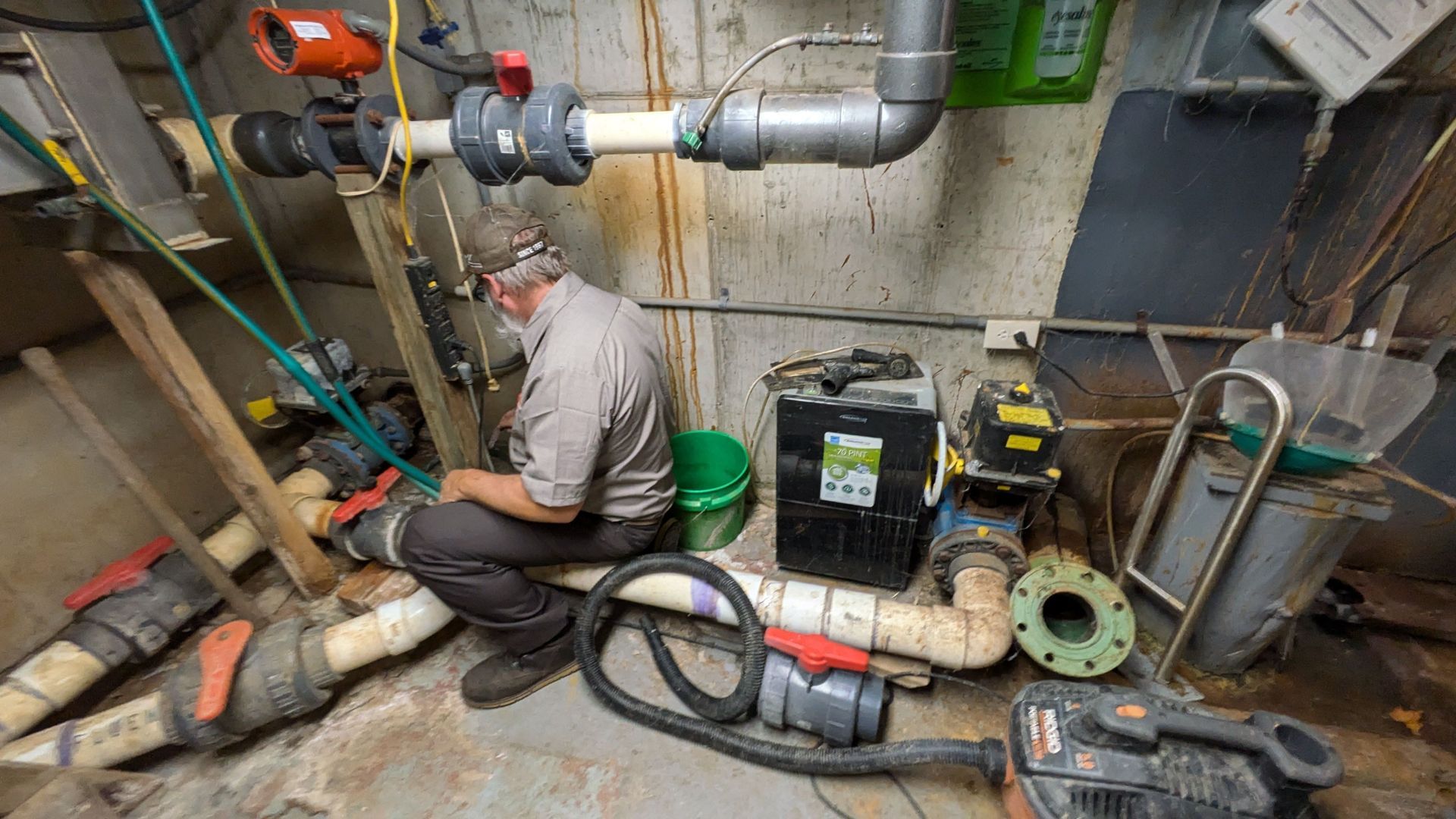 A man in work clothes repairs plumbing in an industrial setting, surrounded by pipes, pumps, and equipment.