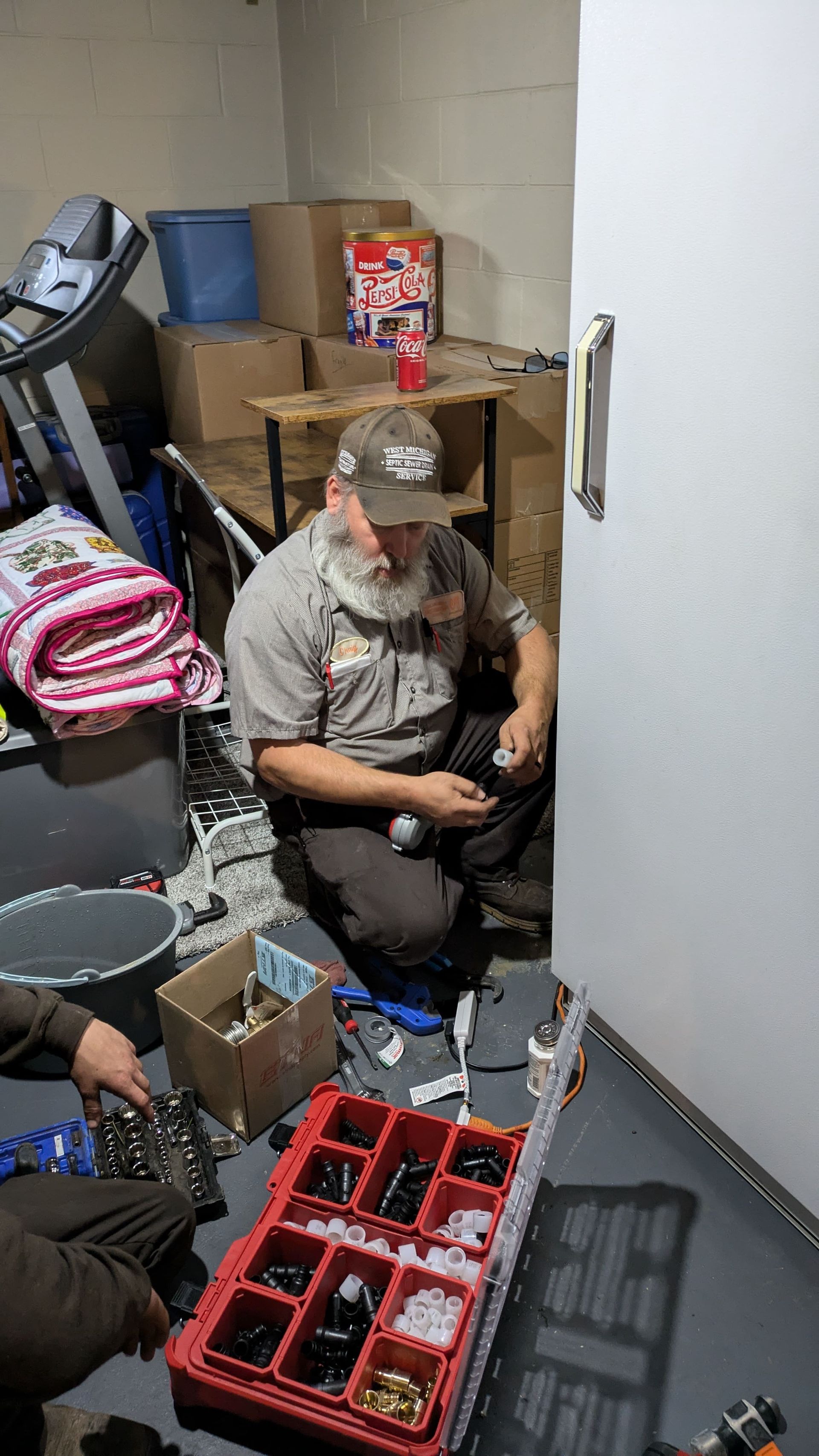 Man in camouflage hat fixing plumbing near a white wall.  Boxes of parts, tools, and a second person are visible.