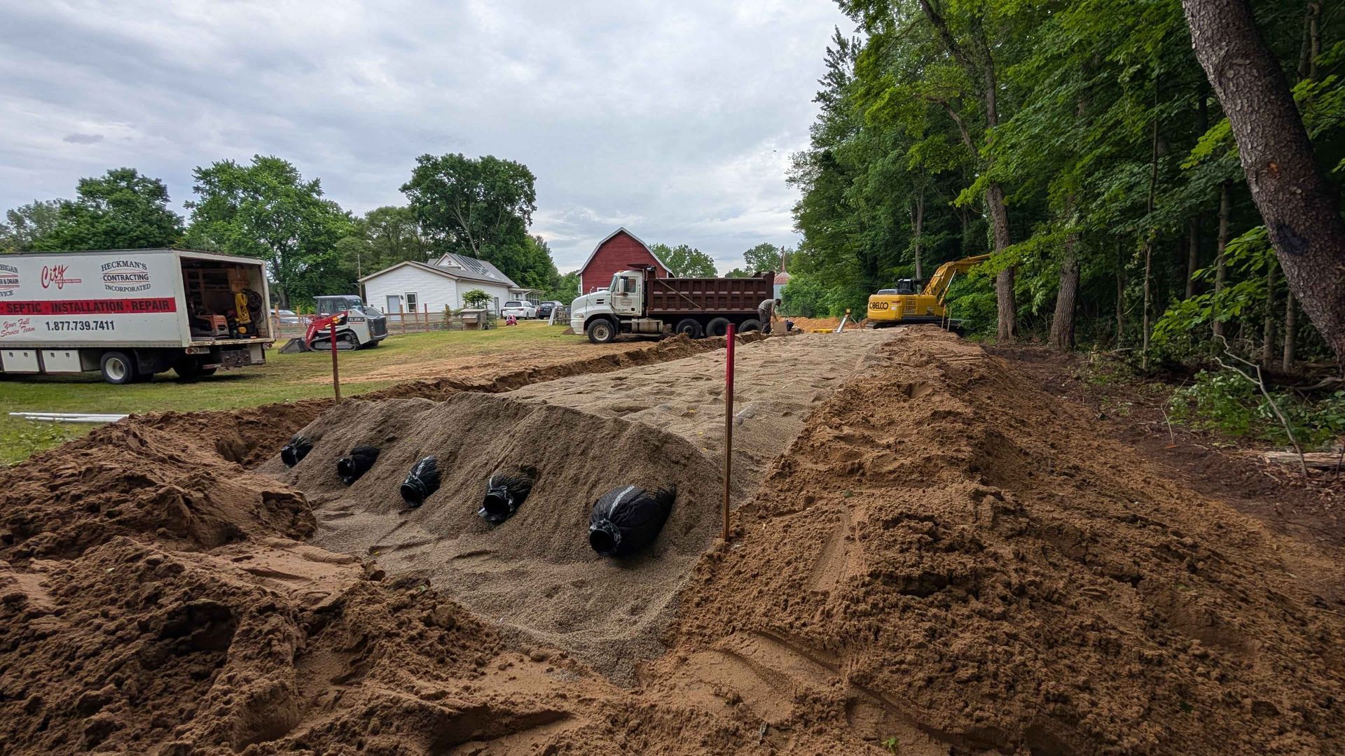Construction site: Excavated ground with black pipes, dirt mounds, trees, trucks, and equipment.
