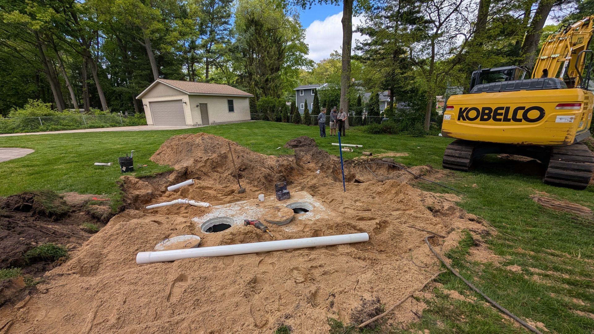 Yellow excavator digging in a yard near a septic system and garage.