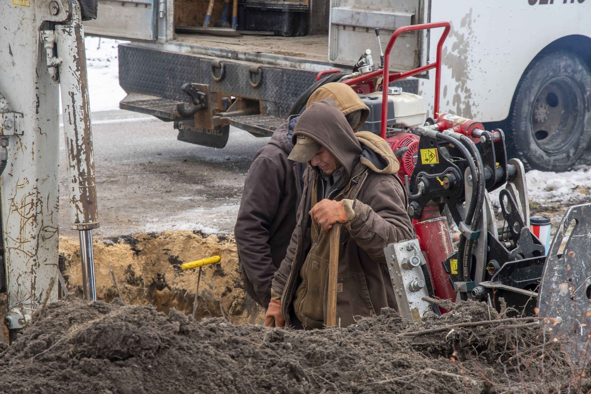 Two workers digging in a muddy trench next to a utility truck on a gray day.