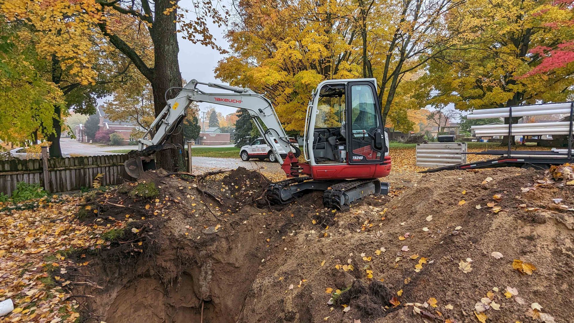 Mini excavator digging a trench in a yard, autumn leaves and colorful trees in the background.