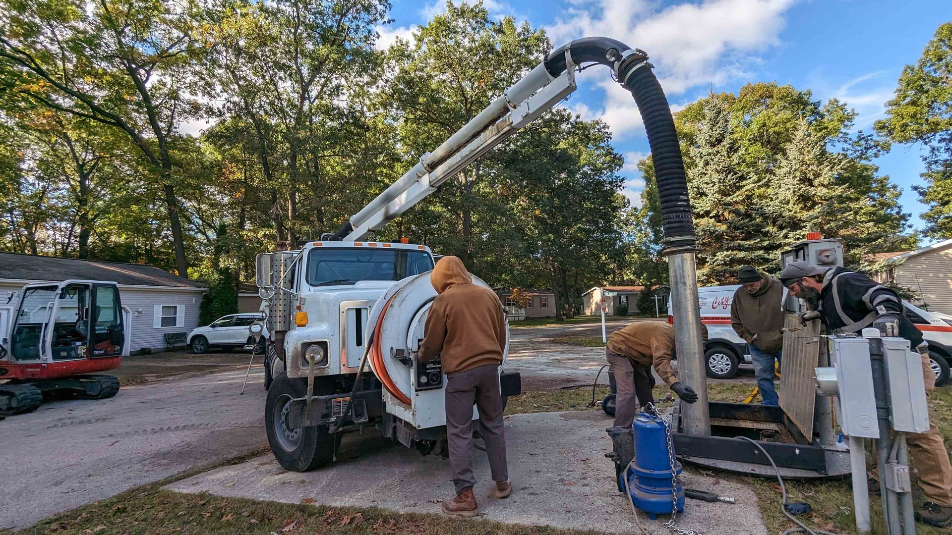 Workers with machinery are servicing a utility near homes, under a cloudy blue sky.