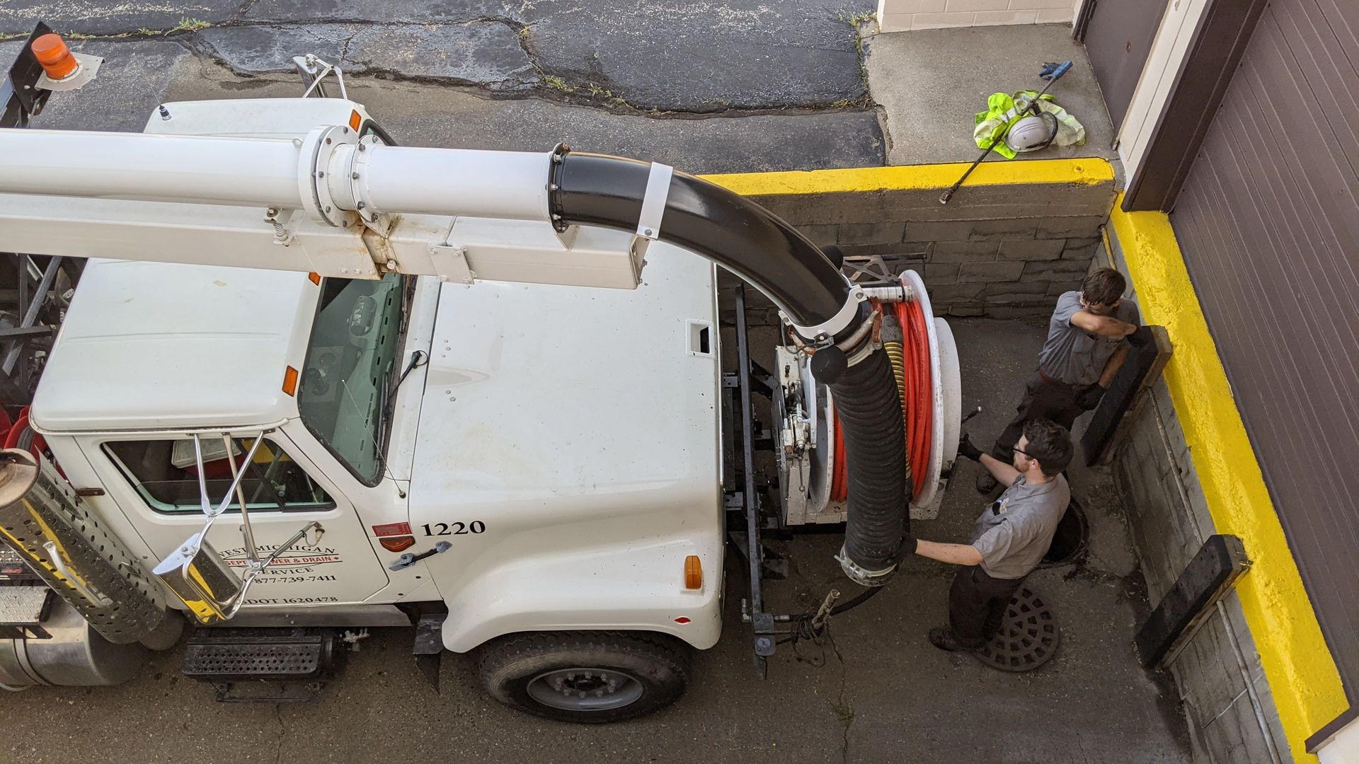 White vacuum truck with a flexible hose reaching toward two workers near a building.