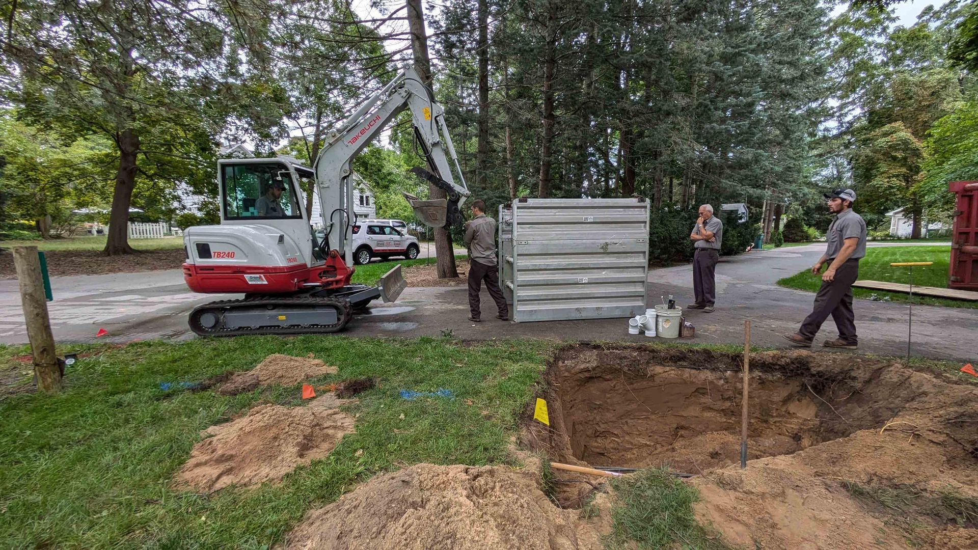 Construction site: Excavator, workers installing a concrete structure near a hole. Green grass, trees, and sky in the background.