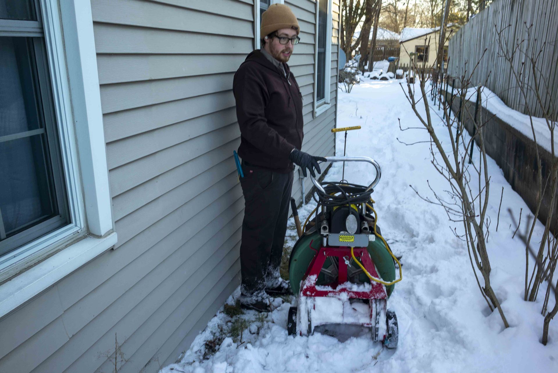 A man in a brown jacket uses a small snowblower to clear snow along a house and fence.