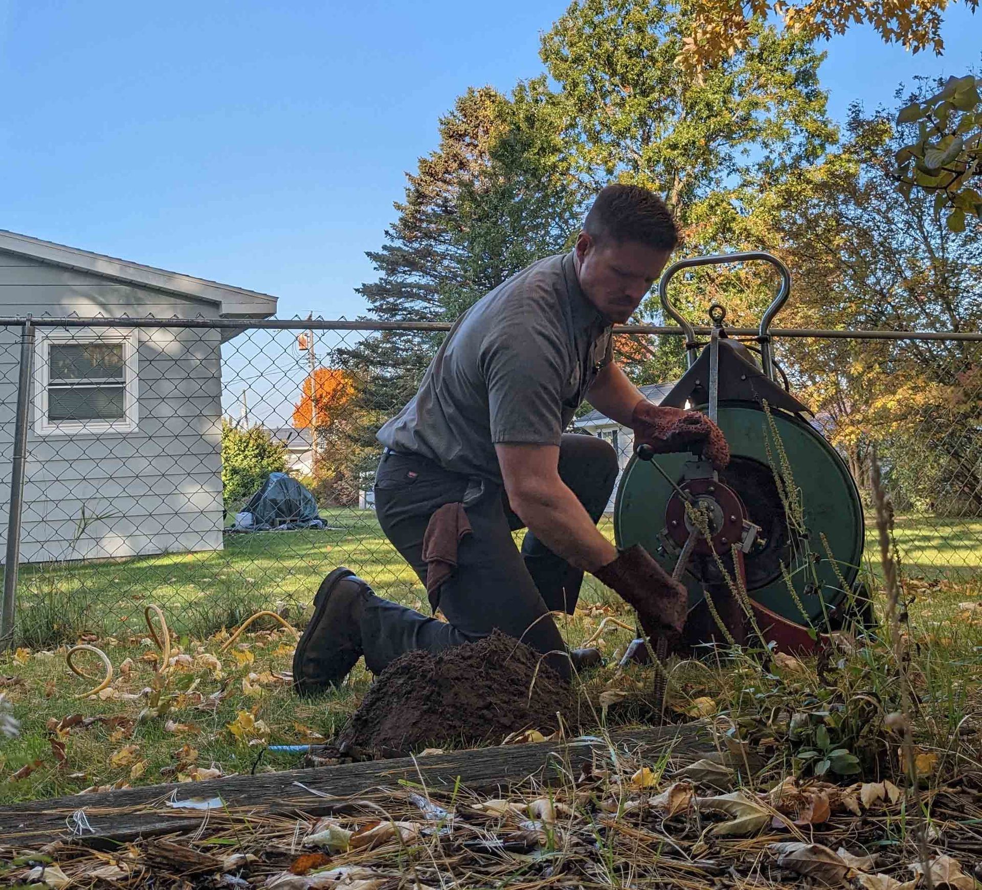 Man kneels in a yard, using a drain snake. Cloudy sky, fall foliage, and chain-link fence in background.