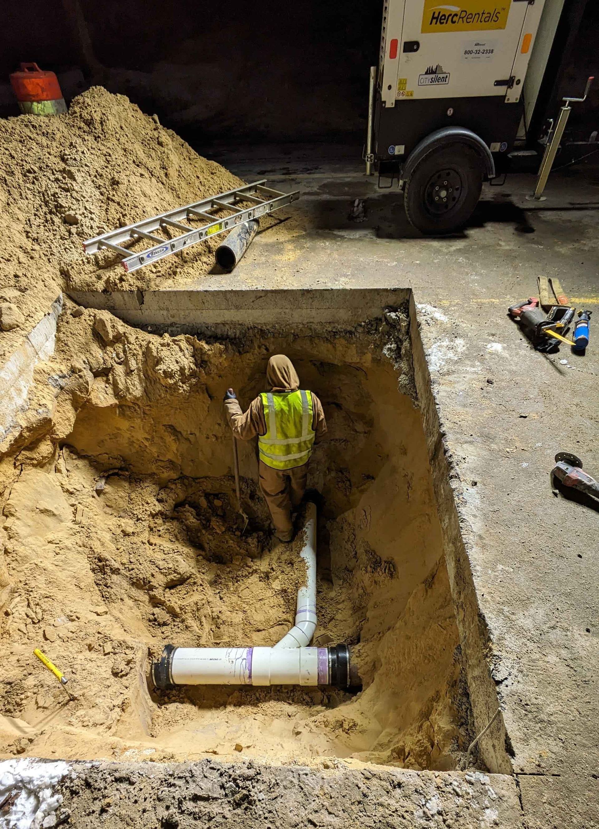 Construction worker in a deep hole, connecting pipes in a concrete area with sand and equipment nearby at night.