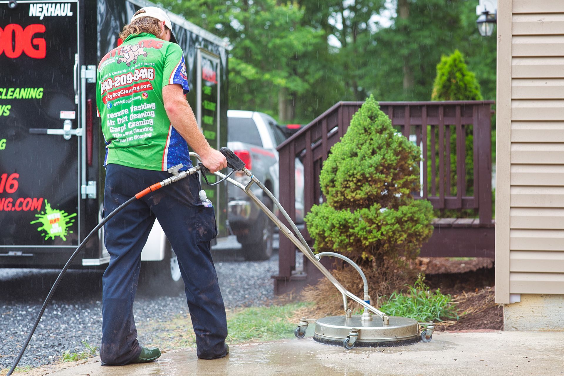Man pressure washing a deck next to a house; black trailer in the background.