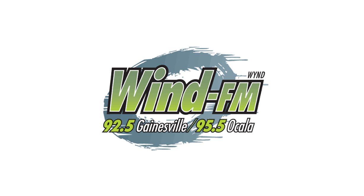Logo for Wind-FM radio, featuring green text on a blue, wind-swept background, with frequencies for Gainesville and Ocala.