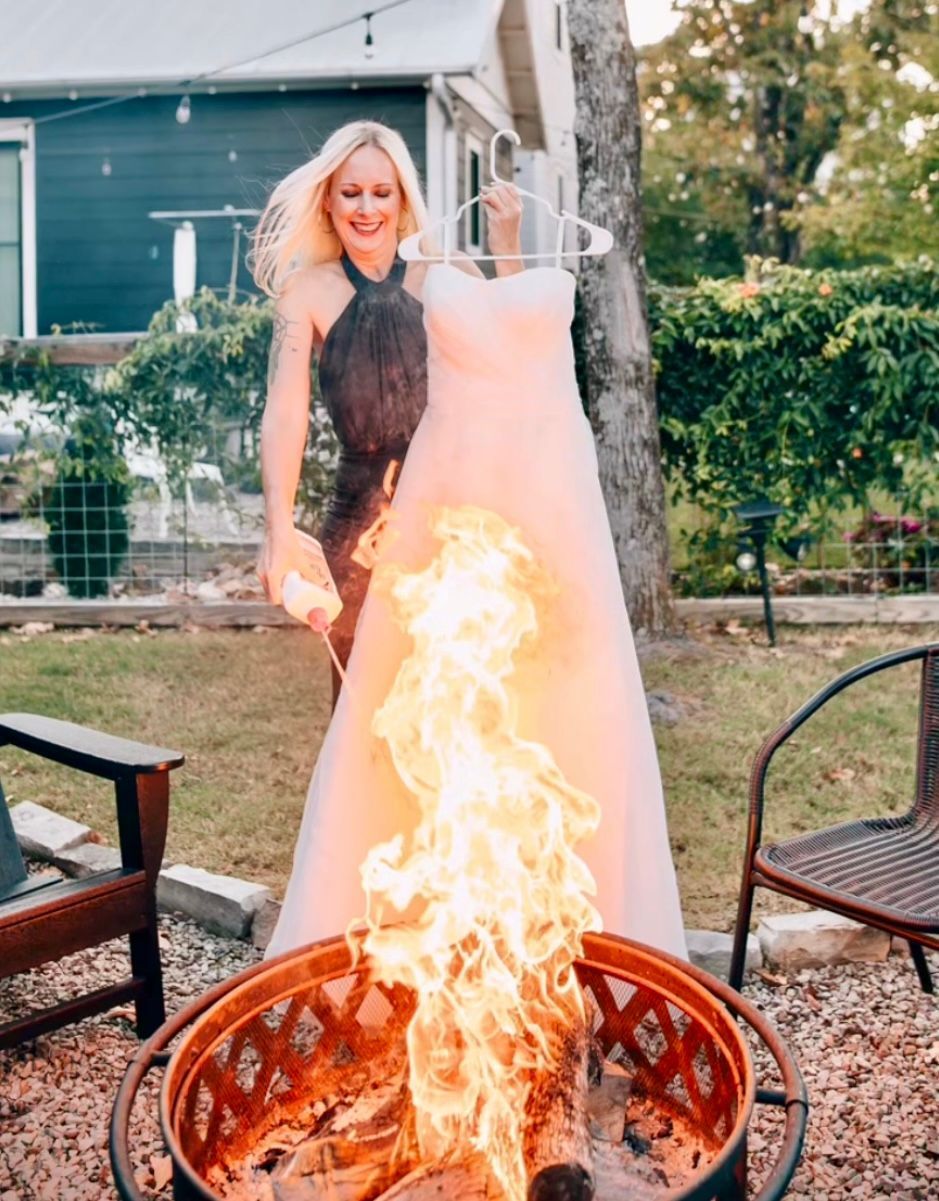 A woman in a wedding dress is standing next to a fire pit holding a hanger.