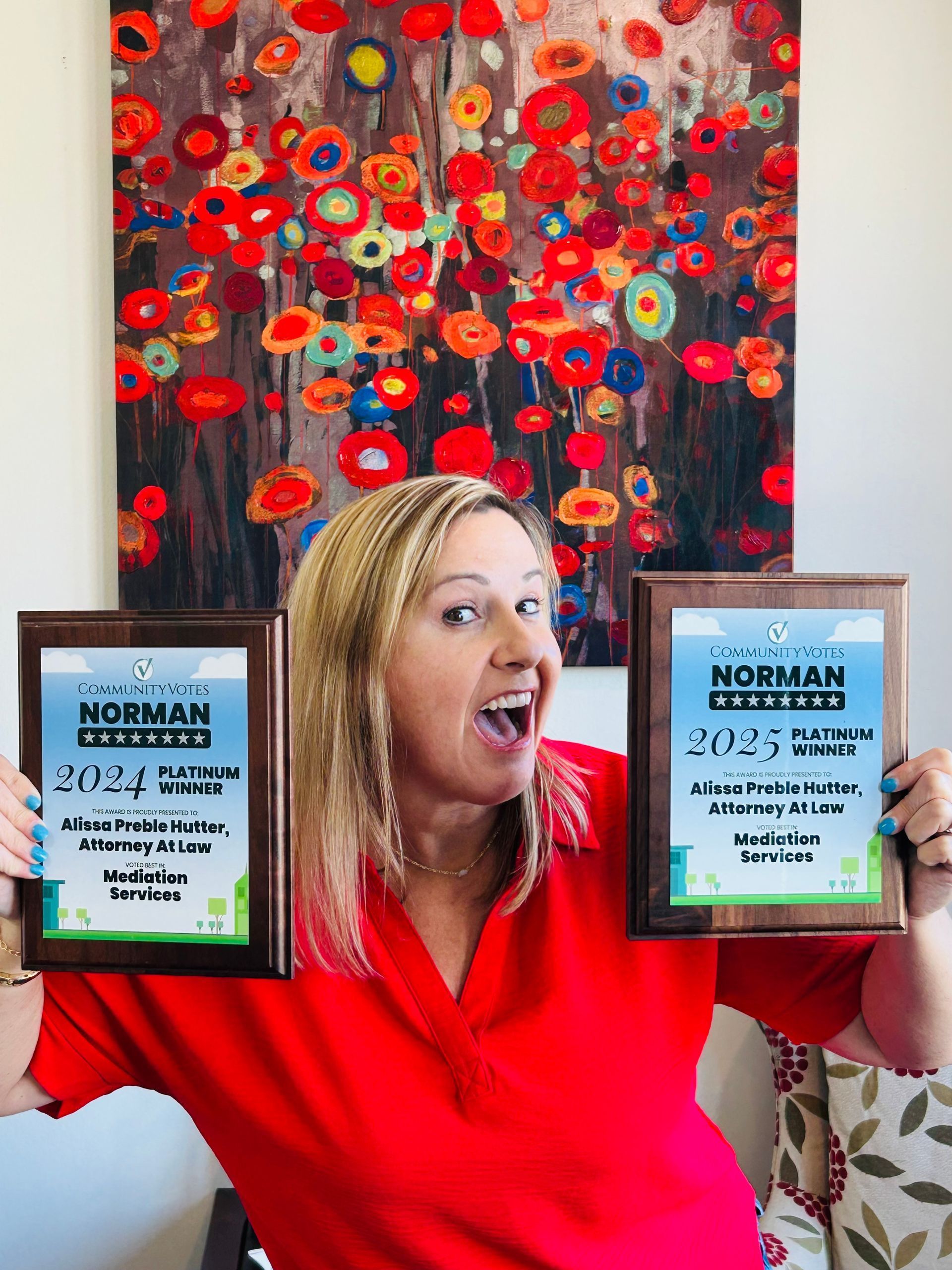 A woman in a red shirt is holding two plaques in front of a painting of flowers.