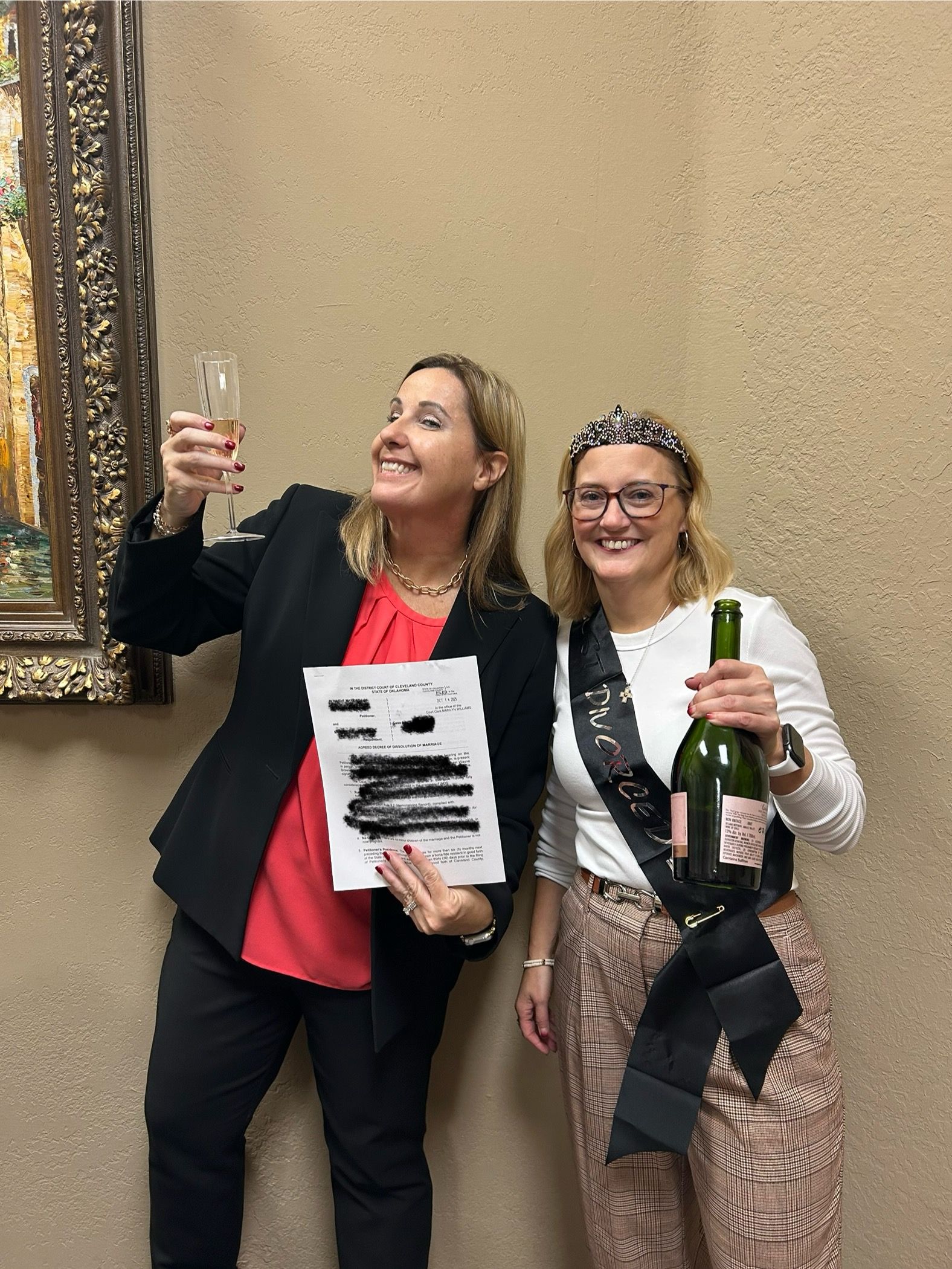 Two women are celebrating; one holds a champagne, the other a document and a glass of champagne. 