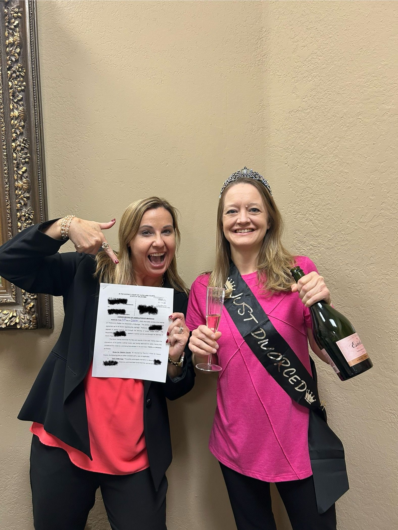 Two smiling women celebrate. One points at a document, the other holds a champagne and a sash that reads “just divorced”.