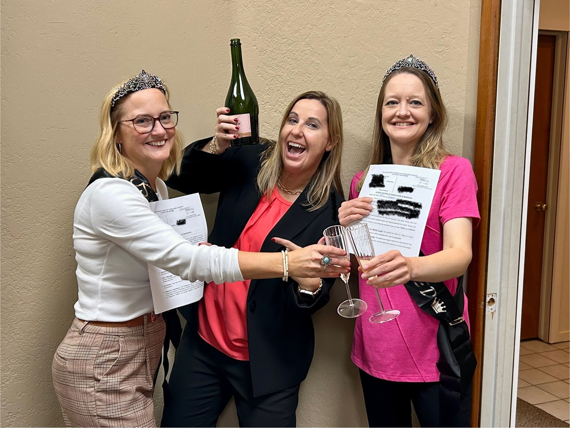 Three women are celebrating with champagne, holding papers, and wearing tiaras in an office setting.