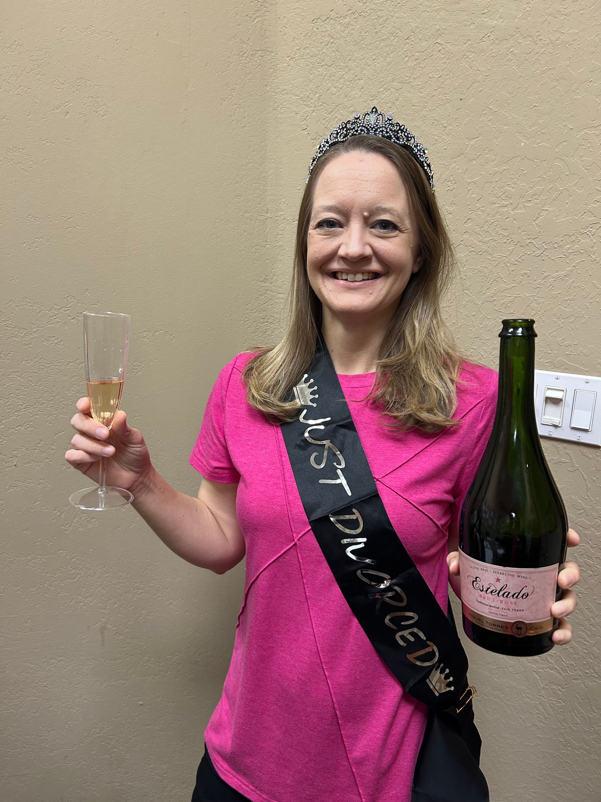 Woman with tiara and sash that says Just Divorced holding a champagne glass and a bottle, smiling.