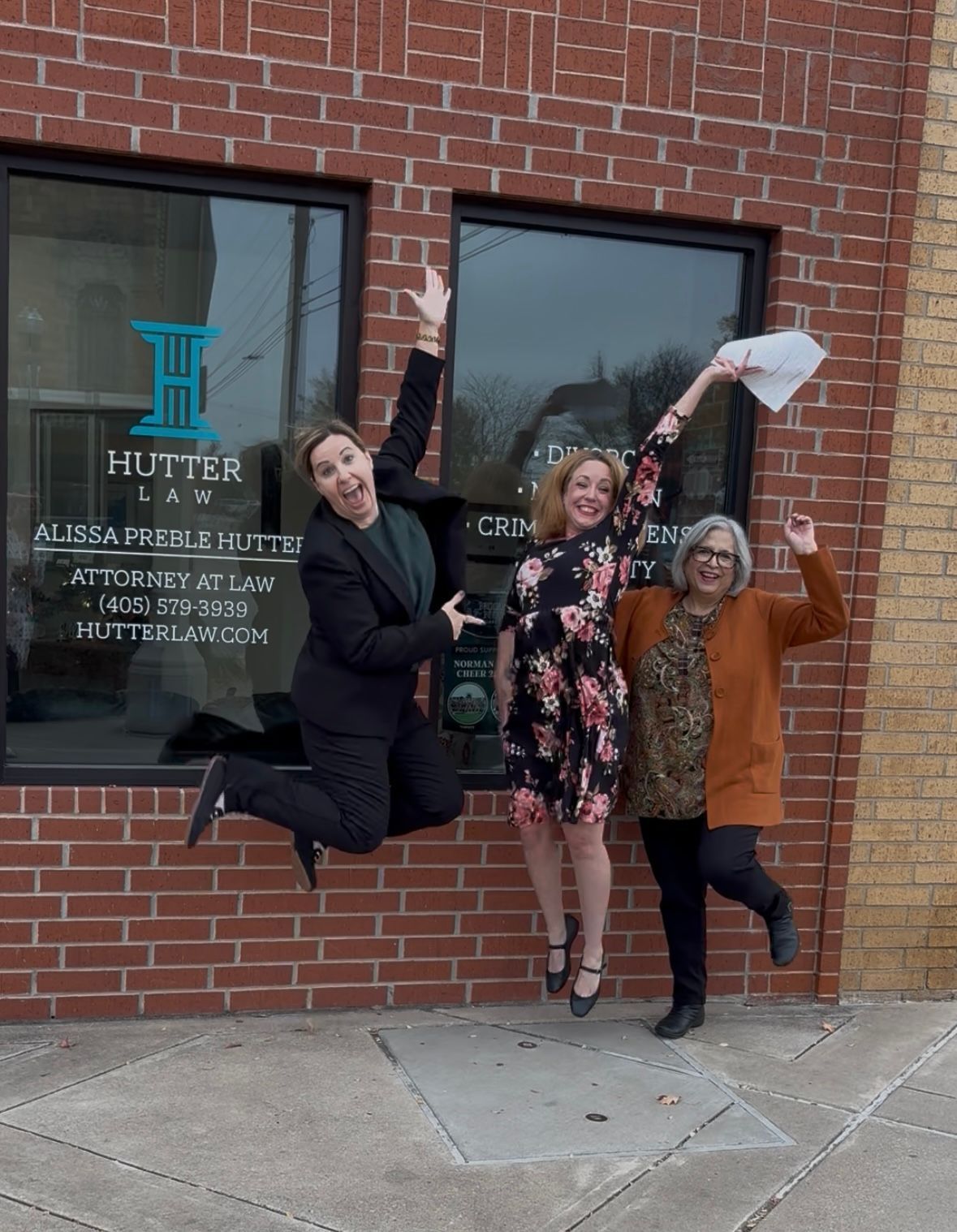 Three people jump for joy in front of Hutter Law building. One holds papers, they smile and celebrate outside.