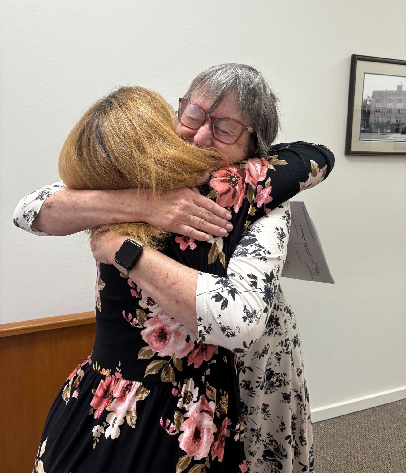 Two women embracing indoors, one in a floral dress, the other in glasses and a similar print dress, both smiling.