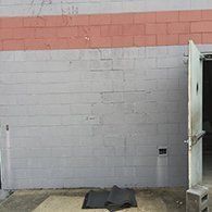 Gray cinder block wall with red brick top; ajar silver door on right, two black mats on the ground.