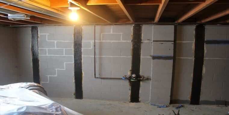 Basement wall with black supports. Gray block walls, exposed ceiling beams, a light fixture, and some debris on the floor.