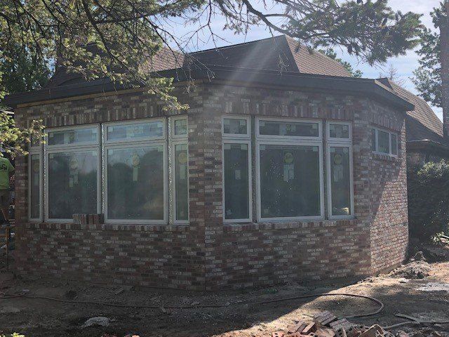 Brick house with large windows under a brown roof, partially obscured by trees.