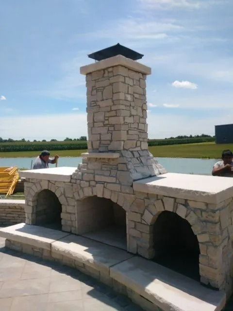 Stone outdoor fireplace with chimney, two arched openings, and two people working in sunny outdoor setting.