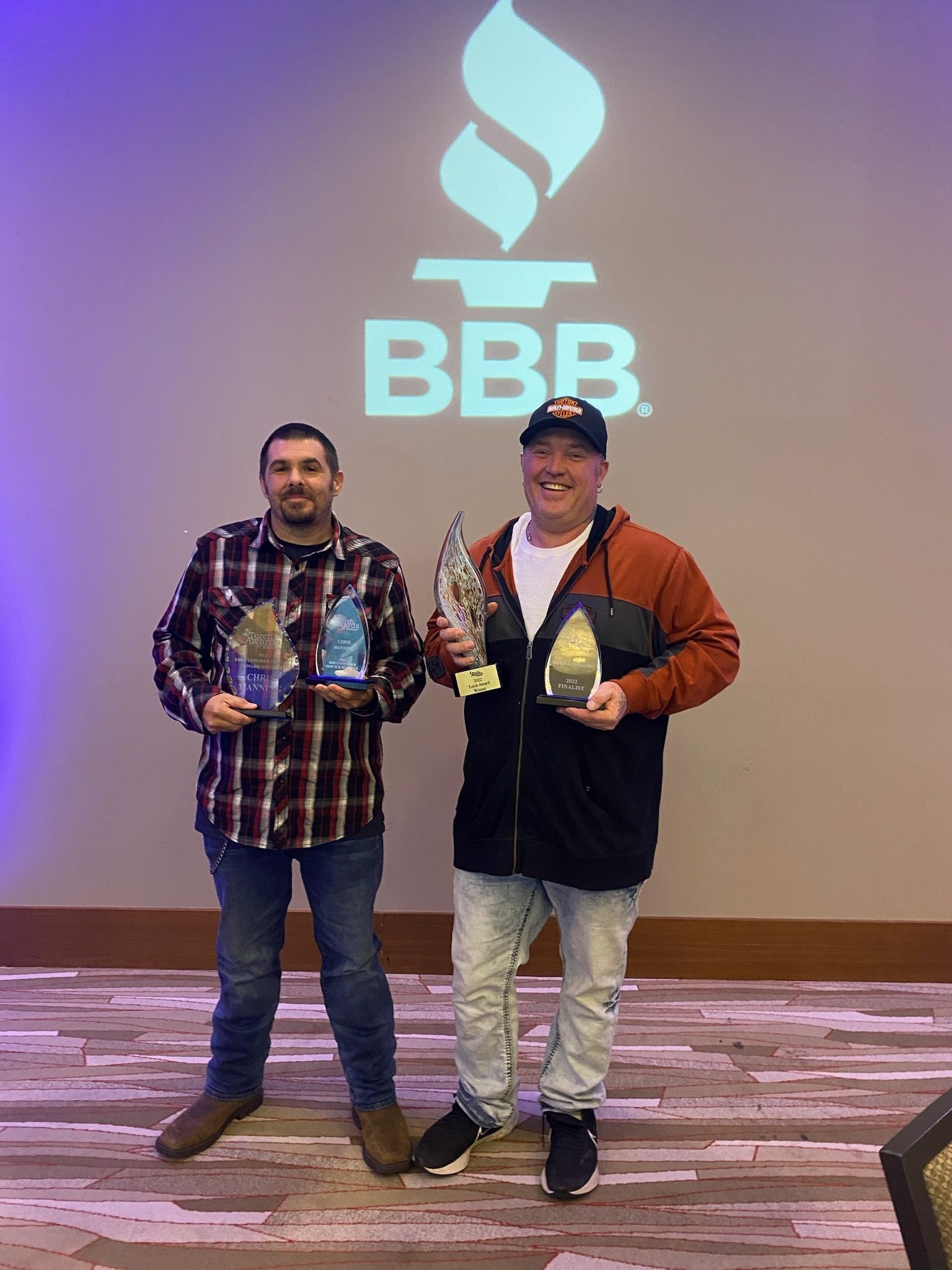 Two men holding awards in front of a Better Business Bureau logo. One wears plaid, the other a jacket.