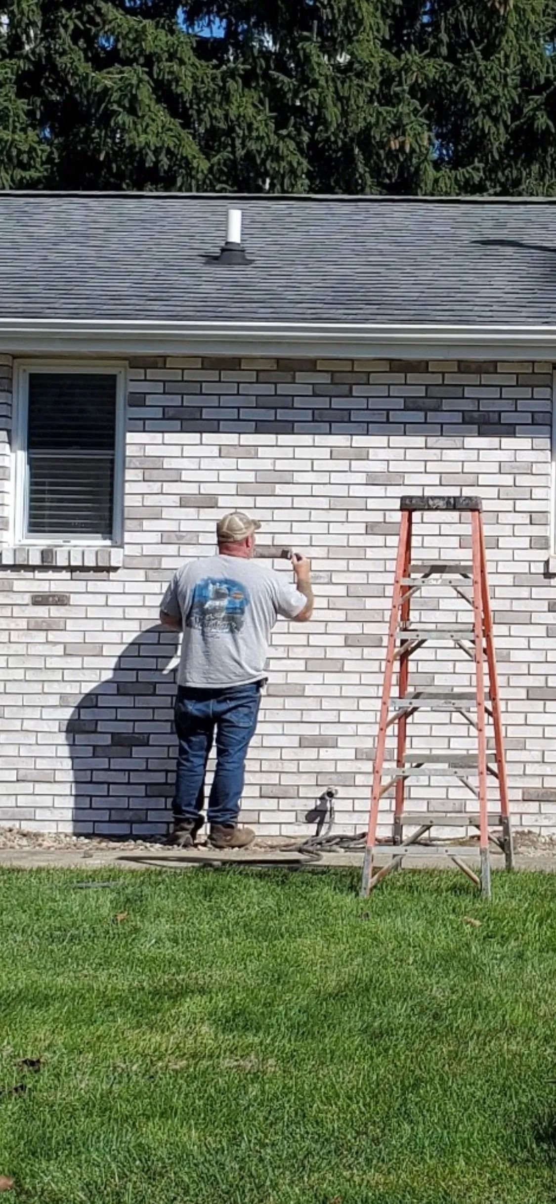 Person painting the exterior brick wall of a house, standing near an orange ladder on a grassy lawn.
