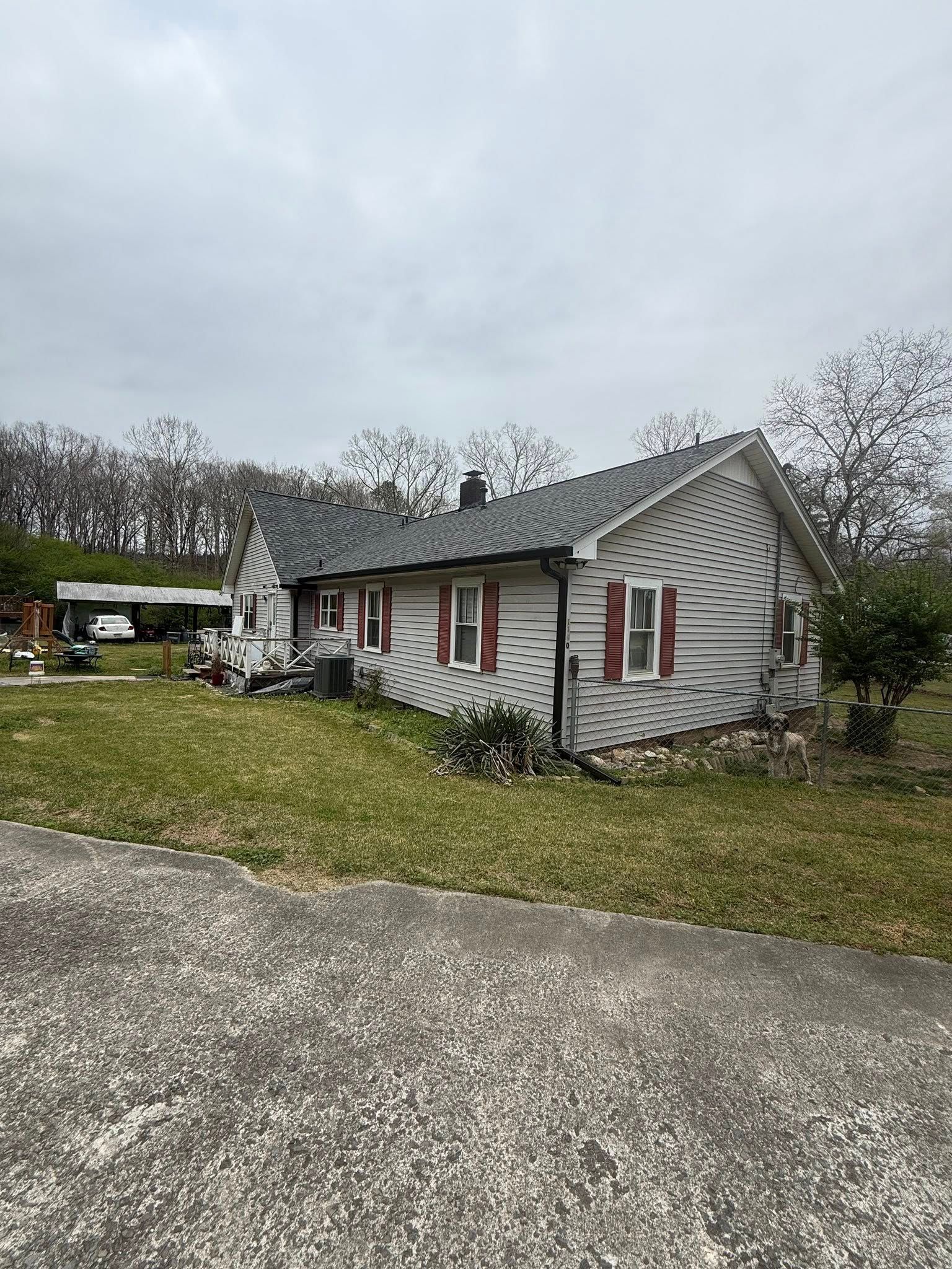 Small white house with red shutters and a warped roof beside a gravel driveway, under cloudy skies