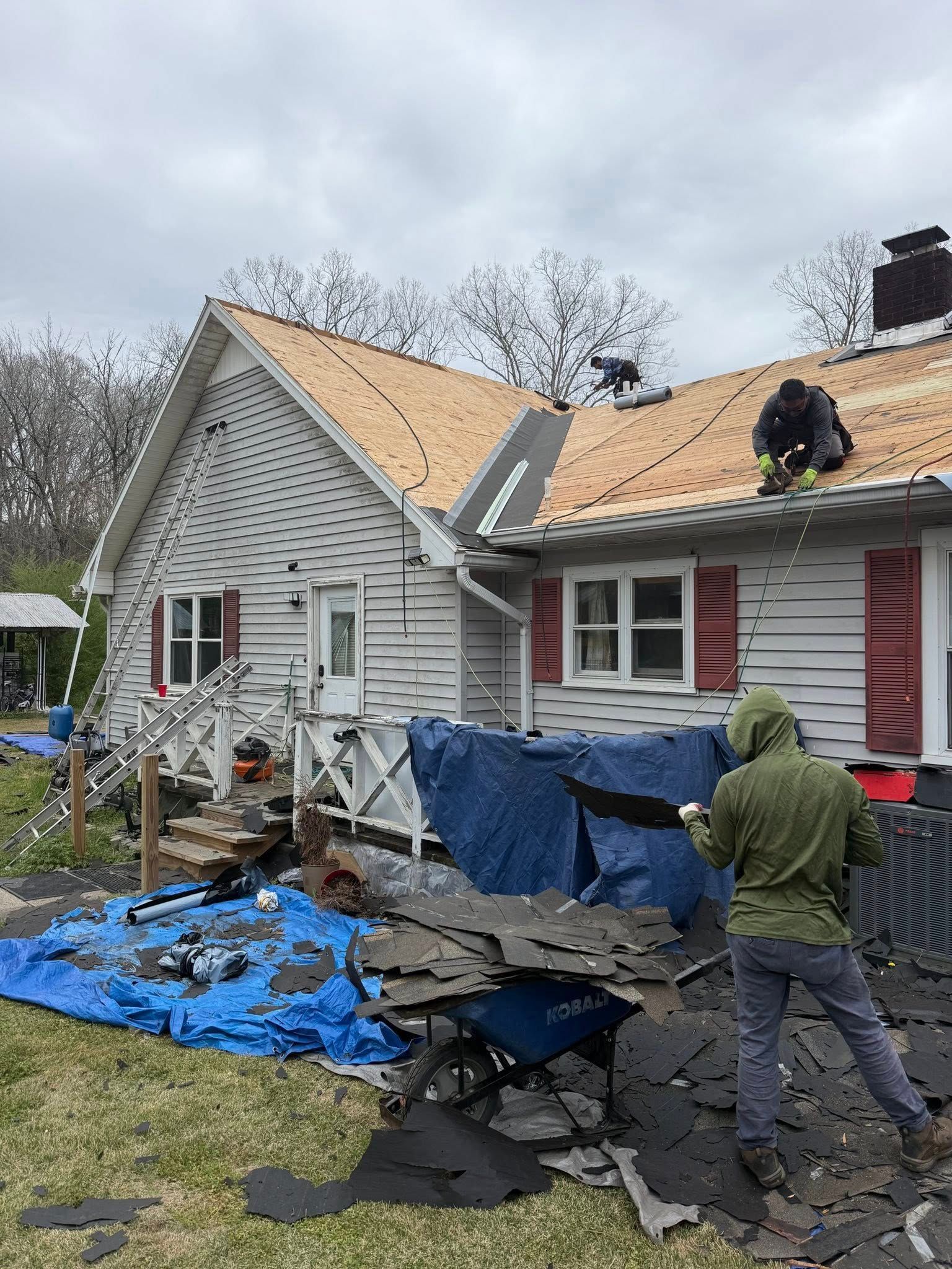 Workers repairing storm-damaged house with exposed roof and blue tarp, debris in front yard