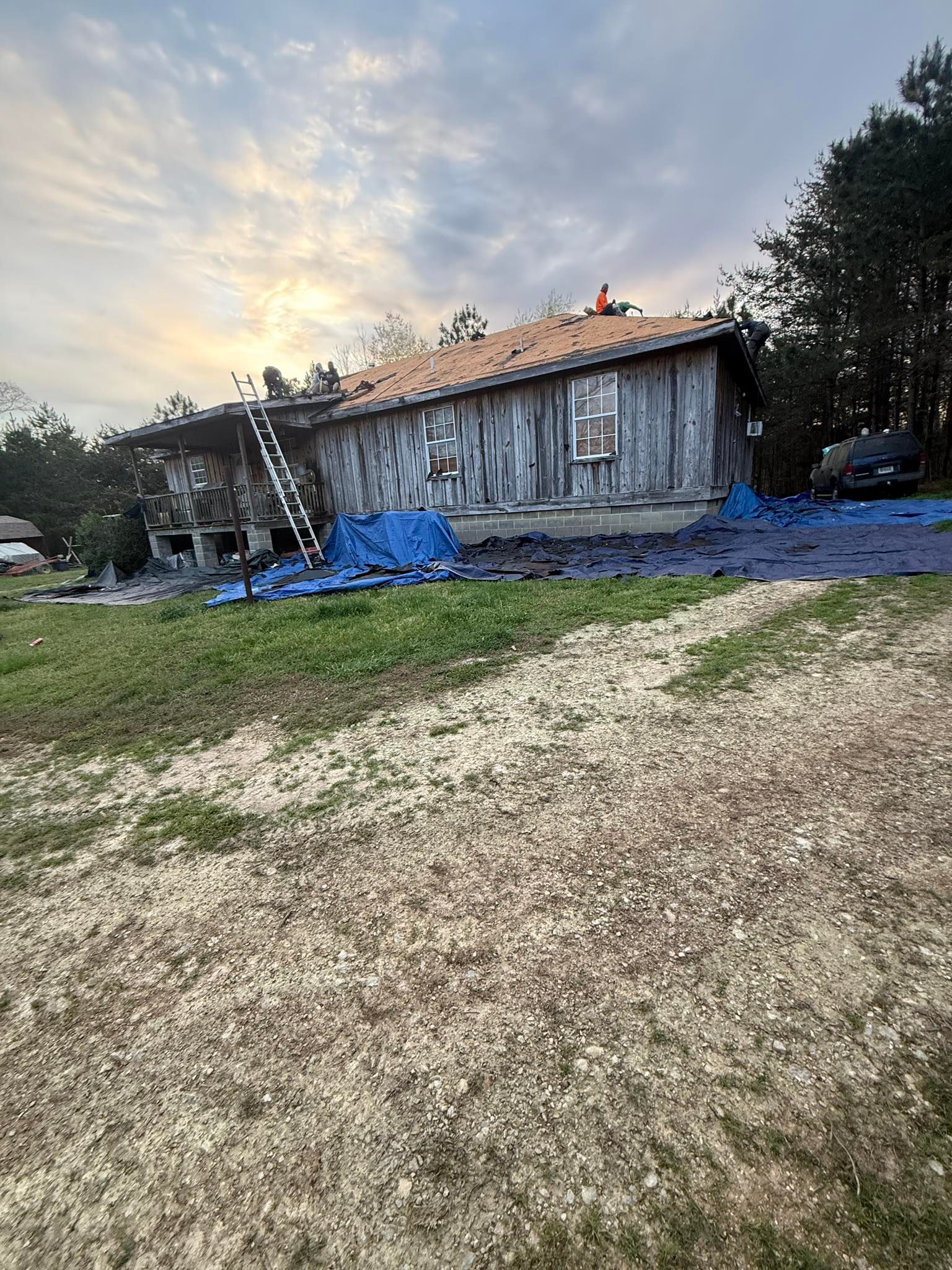 Deteriorating gray wooden house with blue tarp in a grassy yard under a cloudy sunset sky