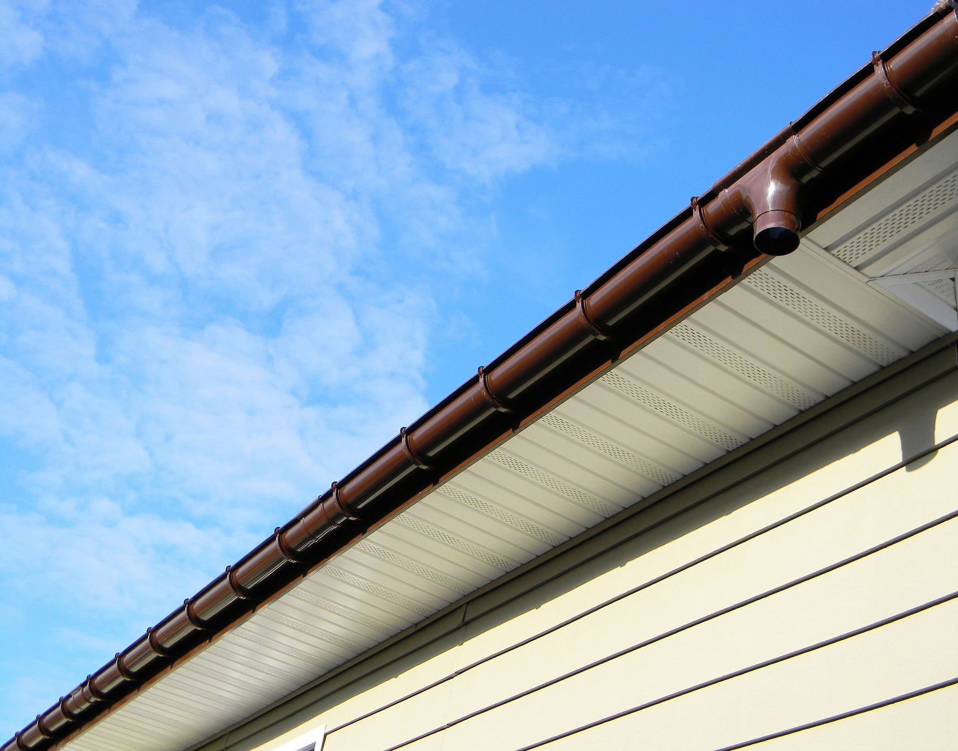 Close-up of a house roof edge with brown gutters against a blue sky.