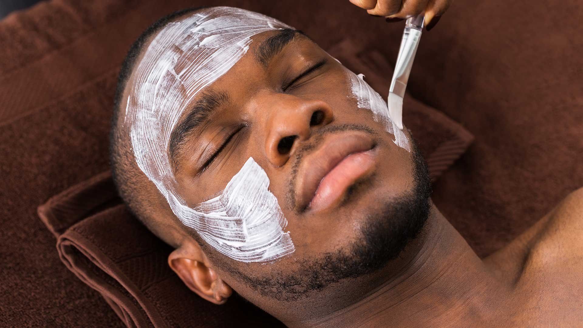Person receiving a white facial mask application while lying on a brown towel