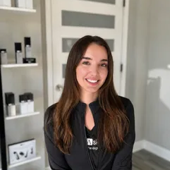Smiling woman in a black jacket standing in a bright room with white shelves and glass panels