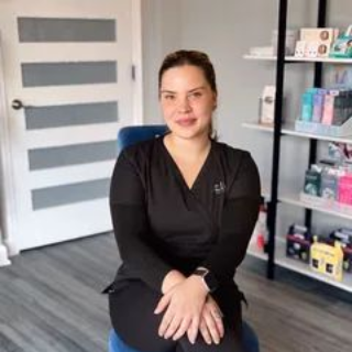 Woman seated on a blue chair in a modern office, wearing black scrubs and smiling at the camera.