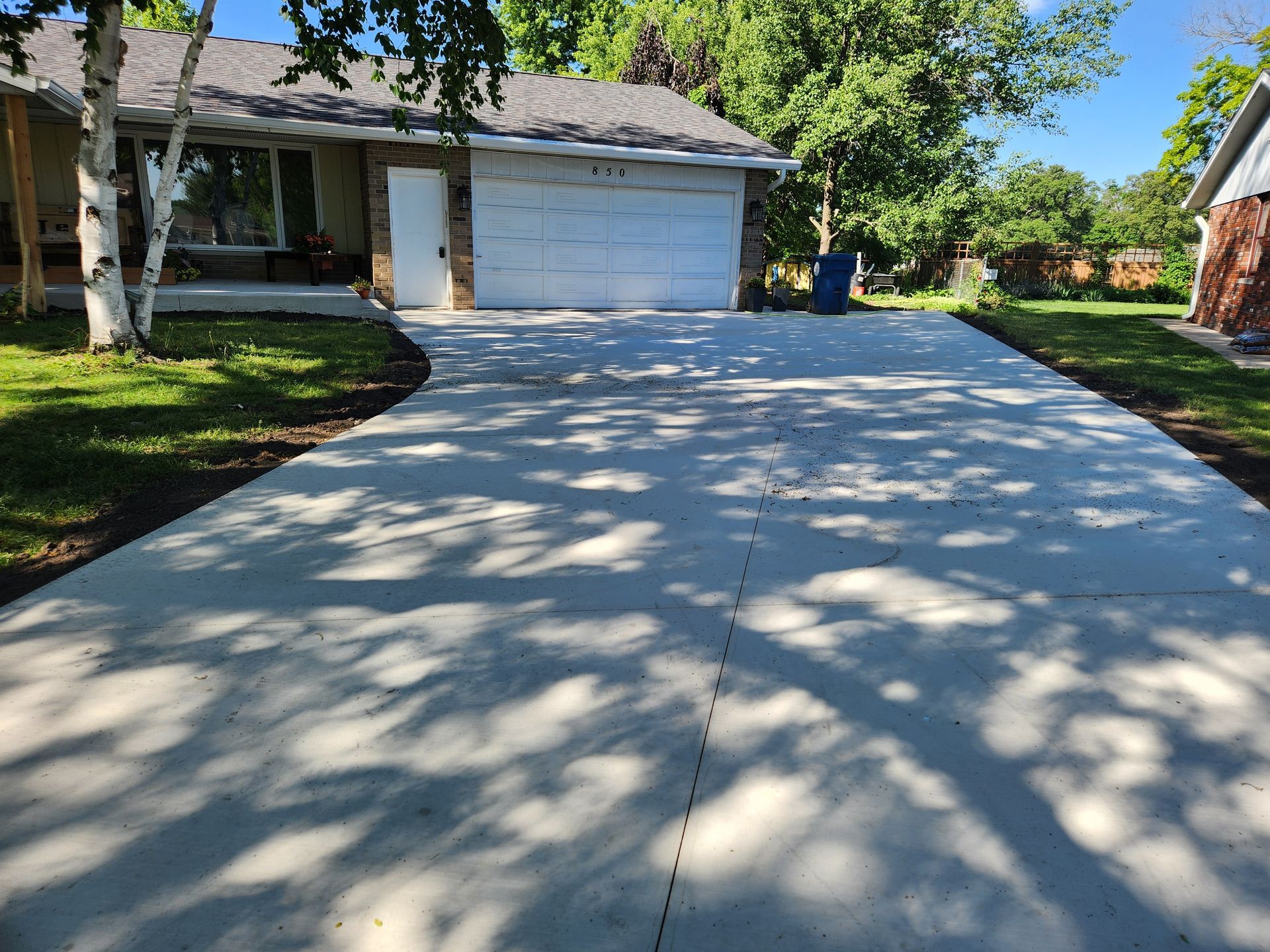Suburban house with a wide concrete driveway, trees casting shadows, and a garage in bright sunlight