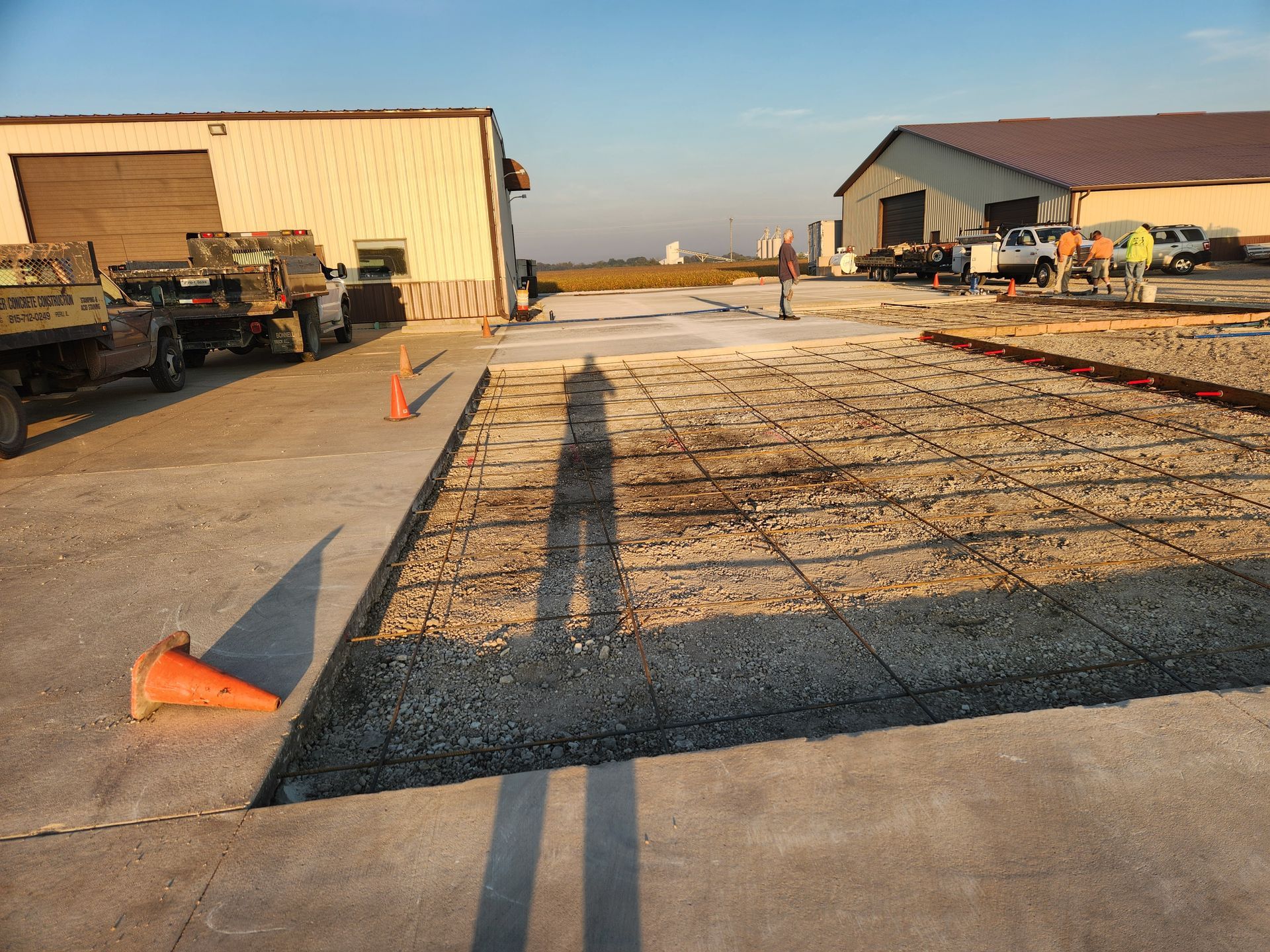 Construction site with concrete work in progress, gravel bed with rebar, two warehouse buildings, and a shadow of a person.