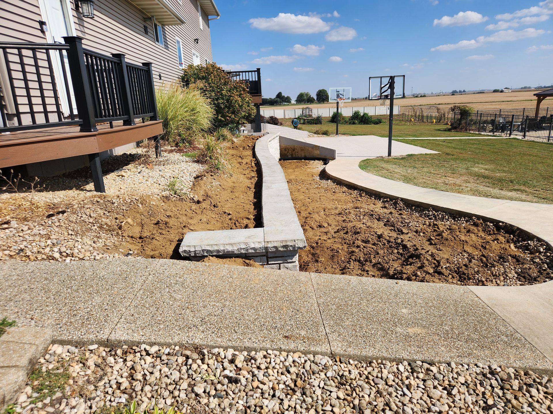 A dirt landscape with a retaining wall, a concrete path, and a deck with black railing under a blue sky.