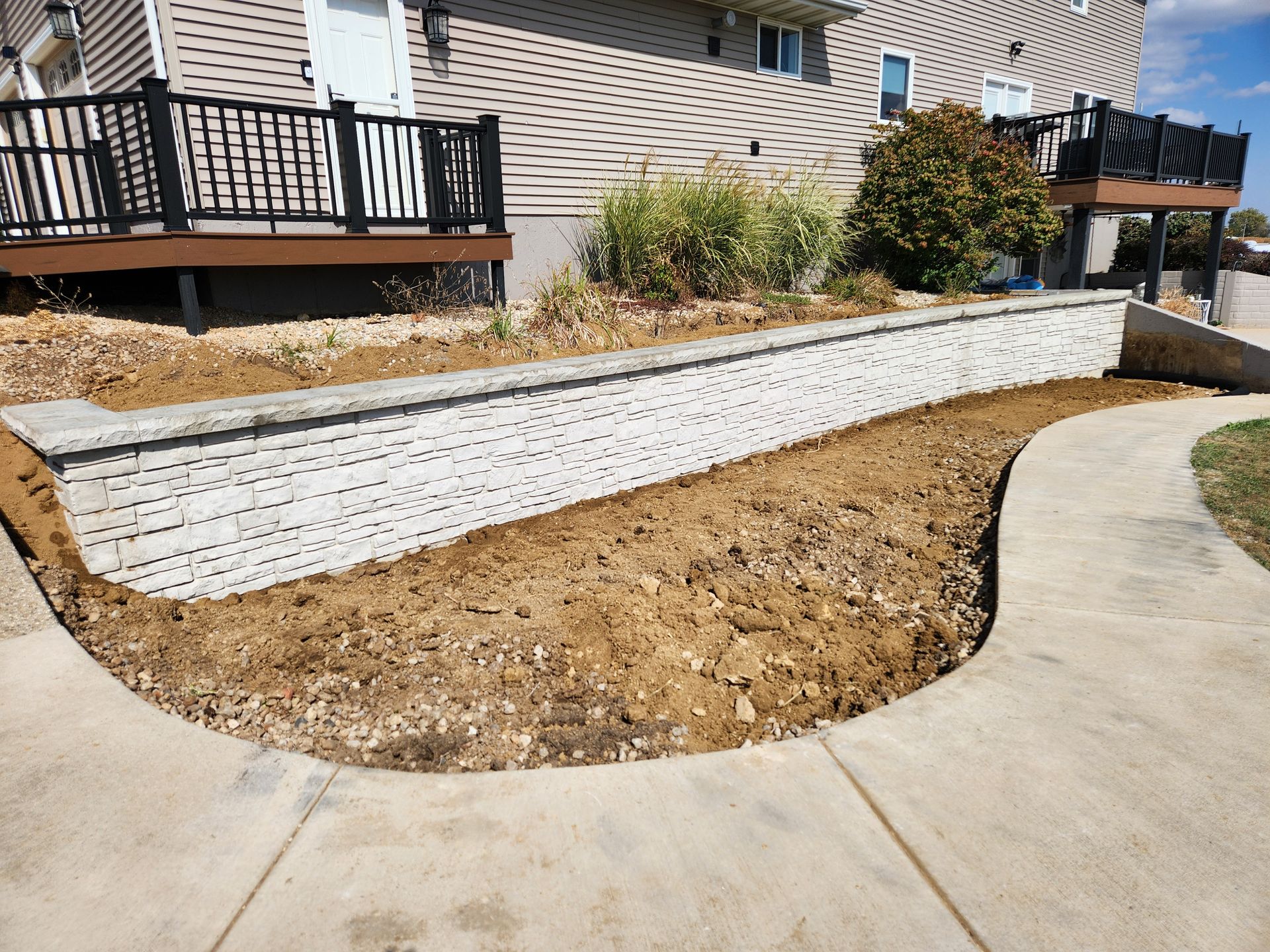 Concrete retaining wall alongside a sidewalk and house with a wooden deck. Brown dirt in front.