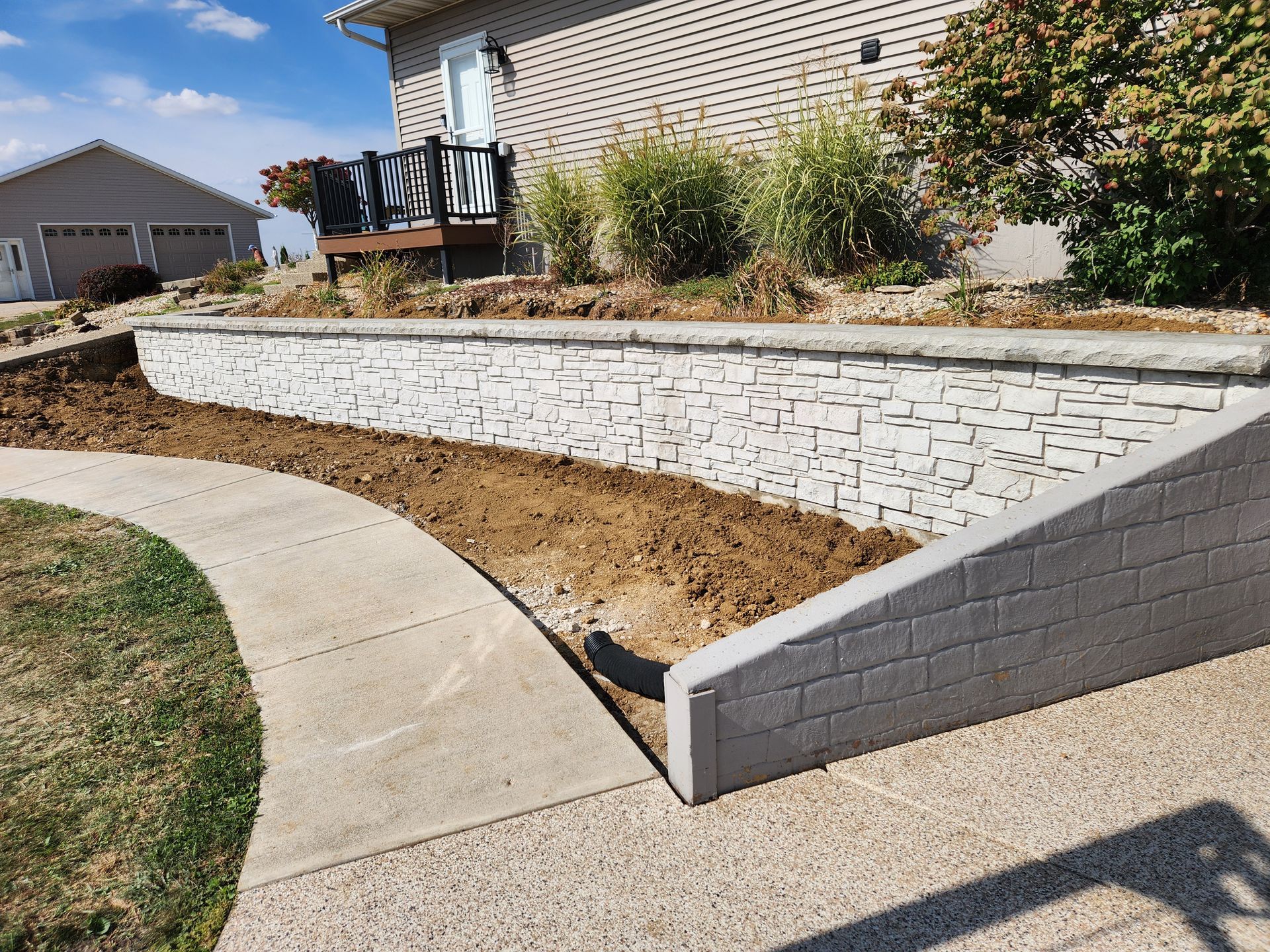 Concrete retaining wall beside a curved walkway with dirt bed for planting. House and garage in background.