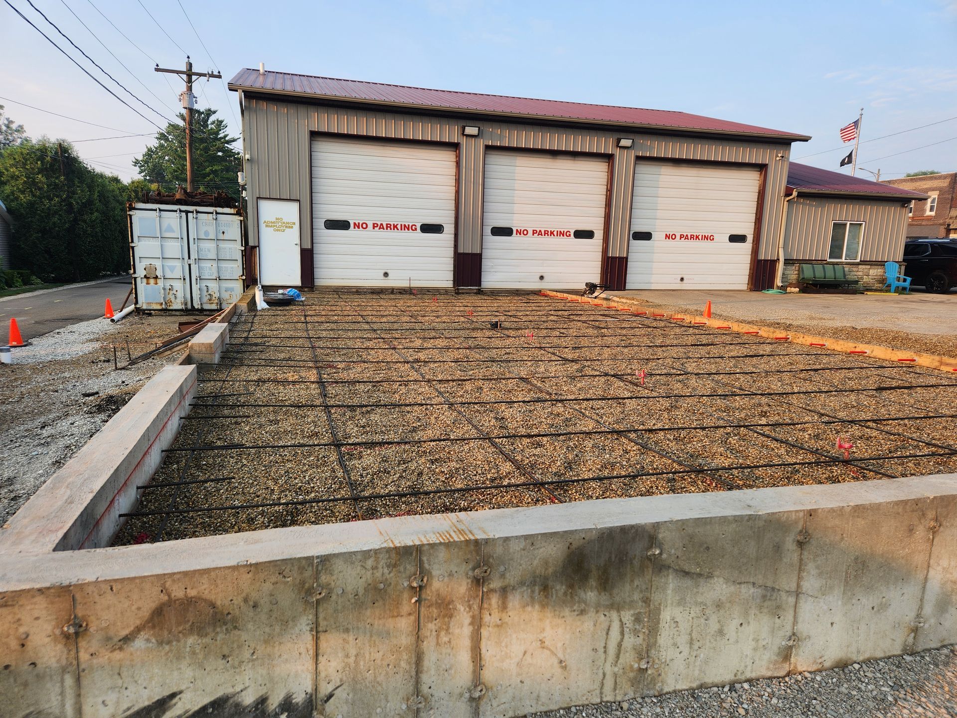 Concrete foundation being prepped in front of a garage with three bays. Rebar grid laid over gravel.