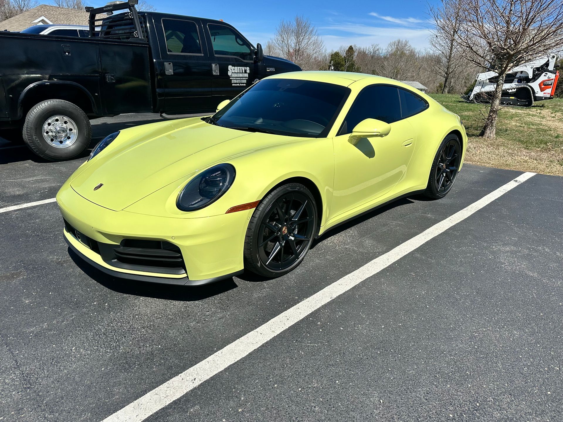Yellow Porsche 911 parked in a lot, black wheels, next to a black truck, sunny day.