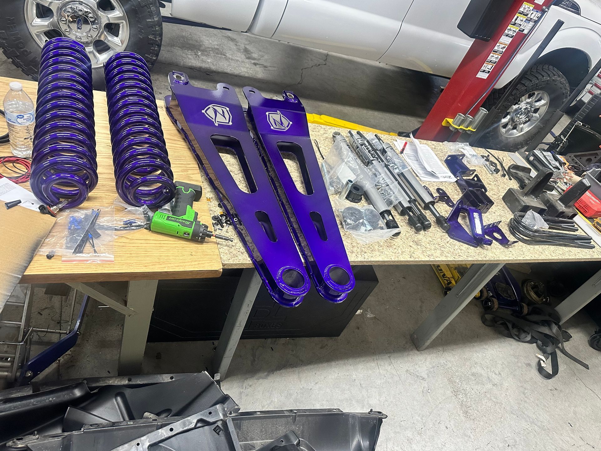 Purple suspension parts and springs on a workbench in a shop, tools and vehicle visible.