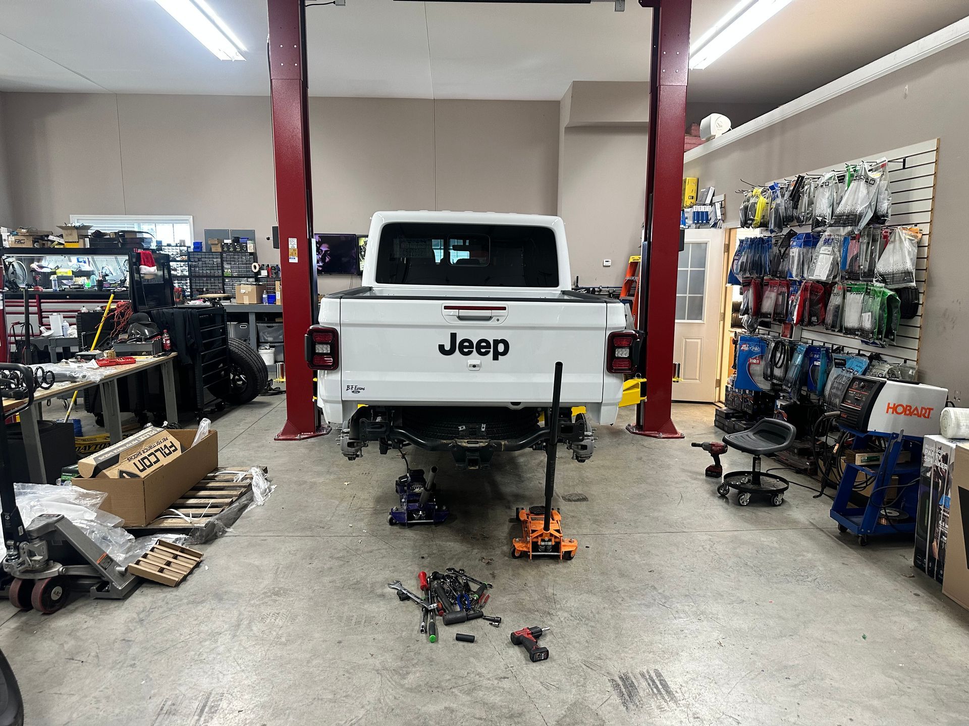 White Jeep Gladiator pickup in a repair shop, rear bumper removed. Tools scattered on the floor.