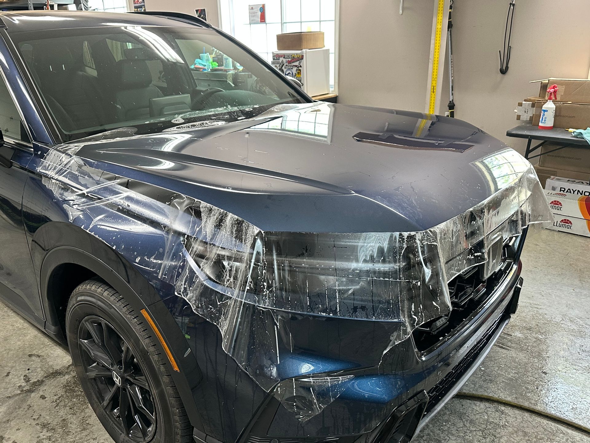 Blue car with protective film applied to the hood and front, in a garage setting.